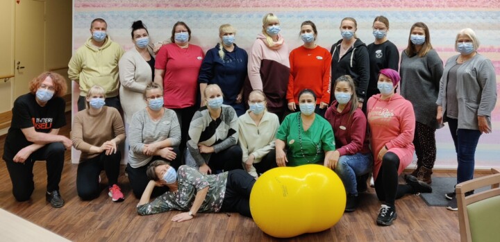 Ryhmäkuva yhdeksästätoista ihmisestä. Kaikilla on maskit kasvoilla. Etualalla yksi henkilö makaa kylkiasennossa ja nojaa kyynärpäähänsä. Hänen edessään on suuri keltainen maapähkinän muotoinen pallo.
